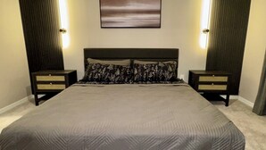 Een groot bed met een gebedruid sprei, twee nachtkastjes met lades, een aan de muur hangend kunstwerk en verticale lampen aan weerszijden van het bed.