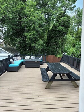 Een houten terras met een zwarte metalen balustrade, een zwarte picknicktafel en een zwarte bank met opdrukken kussens.