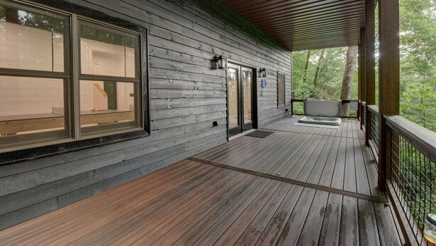 Una terraza de madera con vistas a un bosque, un espacio cubierto con una banca y una puerta con paneles de vidrio.