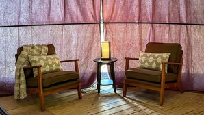 Una habitación con dos sillones de madera, una mesita de noche con una lámpara y un cortinero al fondo.