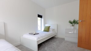 A minimalist bedroom with a single bed, a bedside table, a nightstand, a window, and a door.