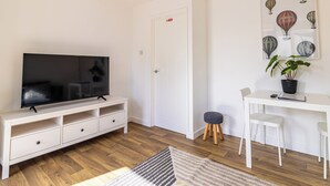 Ein Wohnzimmer mit einem Fernseher auf einem weißen Schrank, einer Tür mit einem roten Schild, einem Bild von Heißluftballons, einer kleinen Pflanze und einem weißen Schreibtisch mit Stuhl.