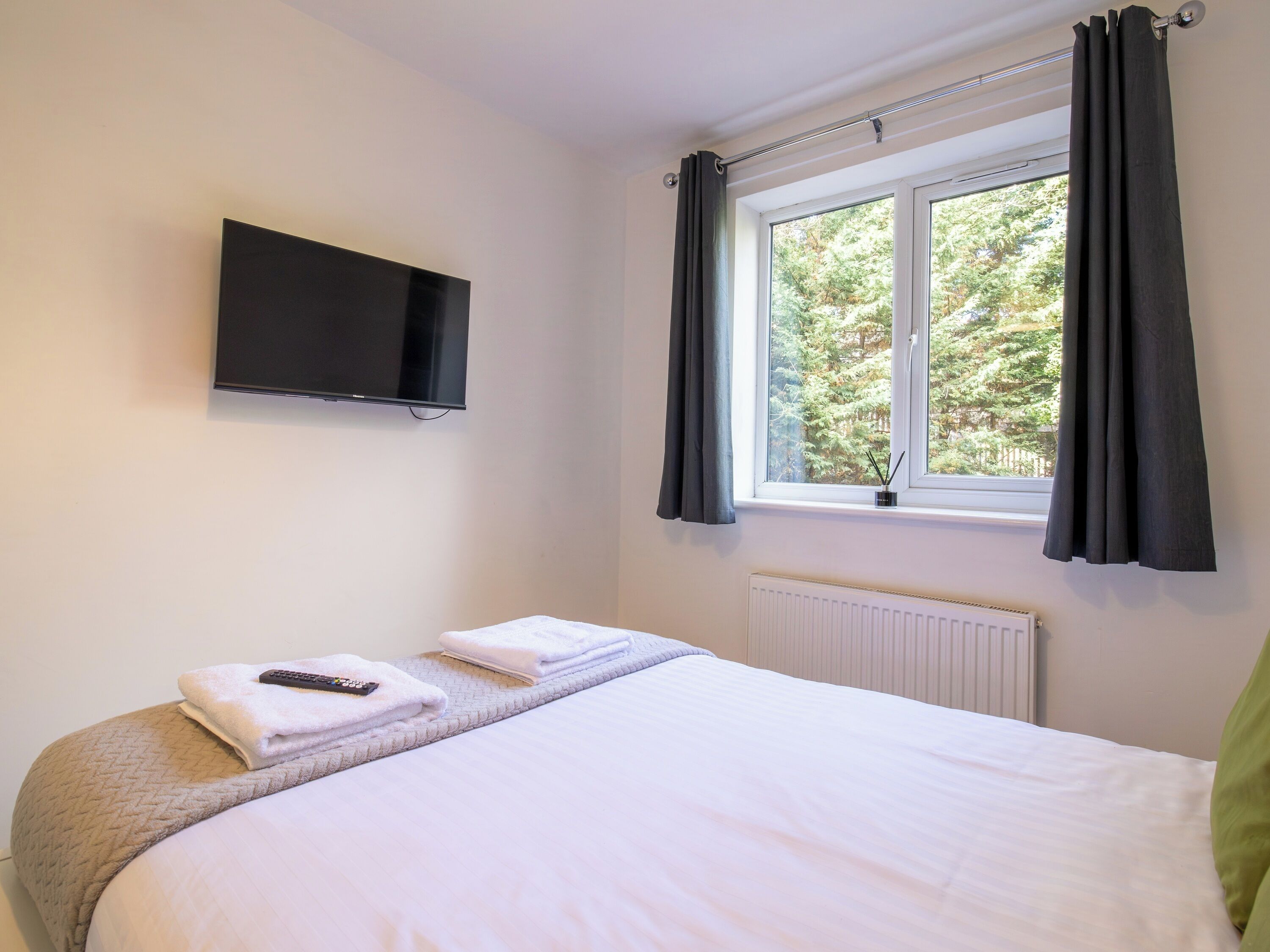 A bedroom with a bed, a TV mounted on the wall, a window with curtains, and a radiator.