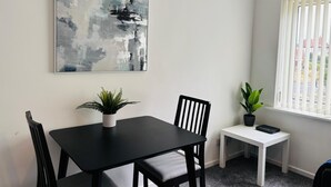 Un comedor con mesa y sillas negras, una mesita auxiliar blanca con una planta, un cuadro abstracto grande en la pared y una ventana con persianas.