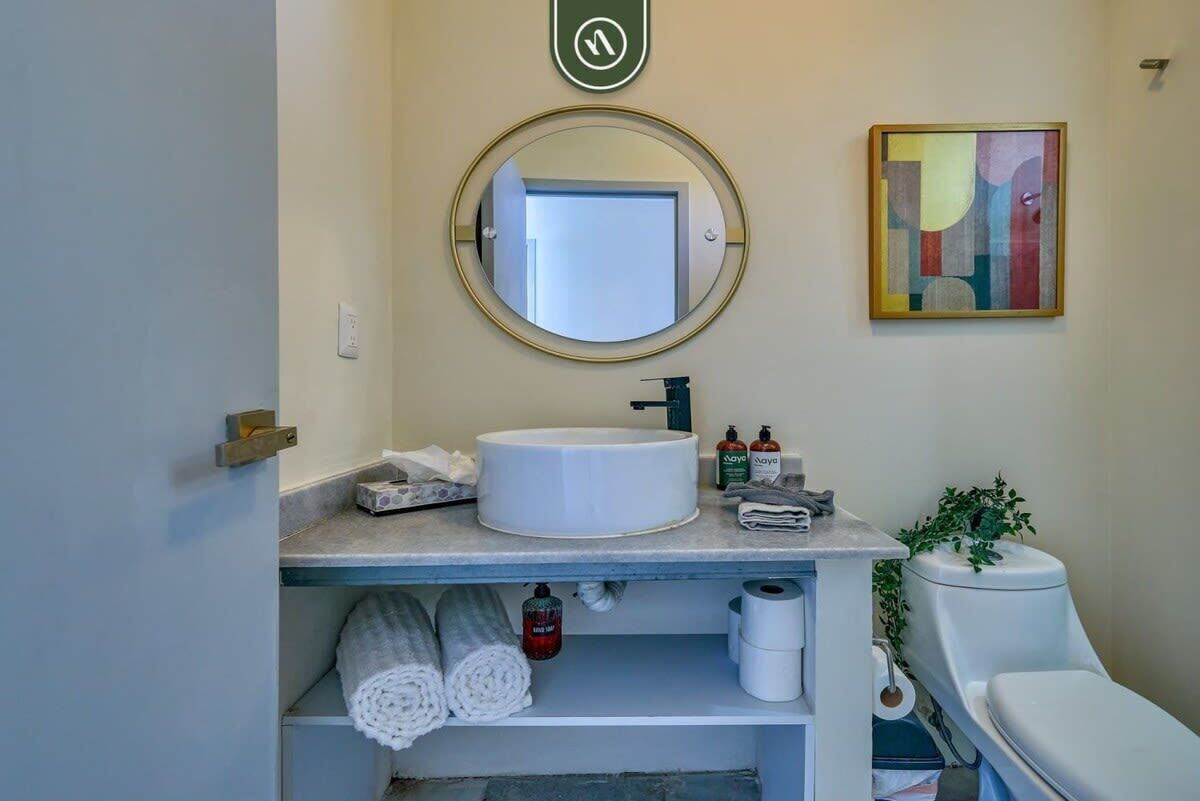 Casa de banho com espelho redondo, lavatório branco e sanita. Há toalhas e produtos de higiene pessoal no balcão.