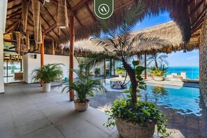 An indoor pool area with a view of the ocean, surrounded by potted plants and a thatched roof structure. - Centric 1 BR Condo with Gym – Amazing Rooftop Pool (Puerto Vallarta)