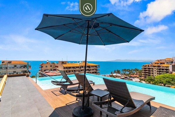 Ett poolområde på taket med solstolar och ett parasoll, med utsikt över ett hotellkomplex med pool och havsutsikt.