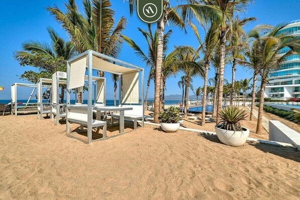 A beach with white lounge chairs and a covered seating area, surrounded by palm trees and a clear blue sky.