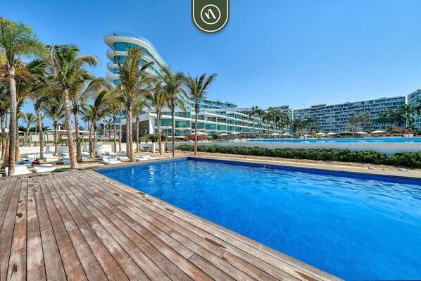 A swimming pool with a wooden deck, palm trees, and a modern hotel building in the background.