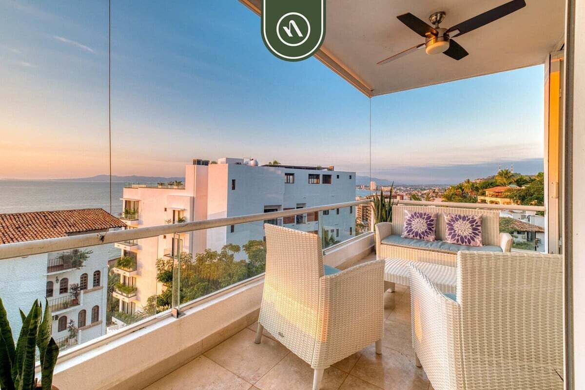 Un balcon doté de meubles en osier, d’un ventilateur de plafond et offrant une vue sur la mer et les bâtiments.