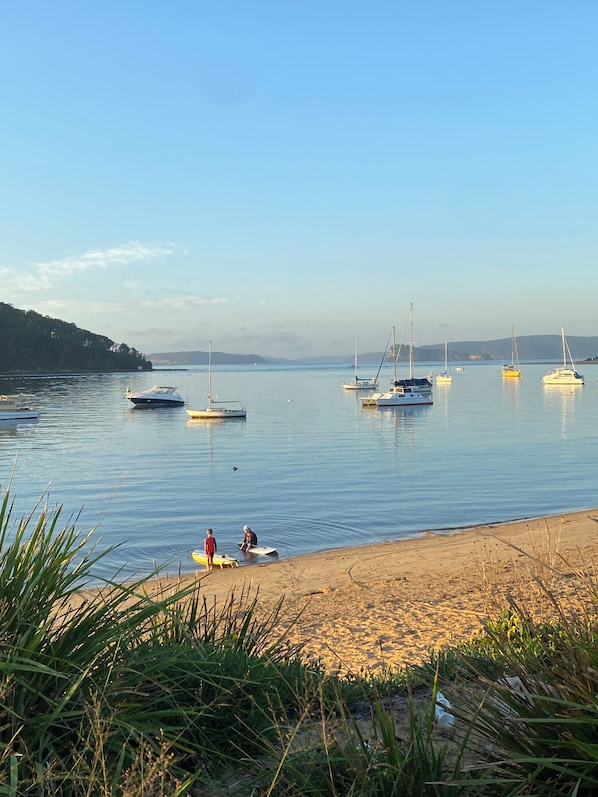 A calm lake with several sailboats anchored. There are two people sitting on the shore, and a sandy beach with grass in the foreground.