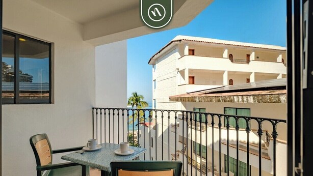 A balcony with a table and chairs, a view of a building with balconies, and a clear sky.