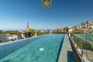 A rooftop infinity pool with clear blue water, surrounded by a metal railing and a view of a coastal town with buildings and a lighthouse in the distance.