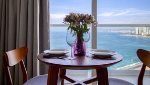 A dining table with plates and wine glasses, a purple vase with flowers, and a view of the ocean and city skyline.