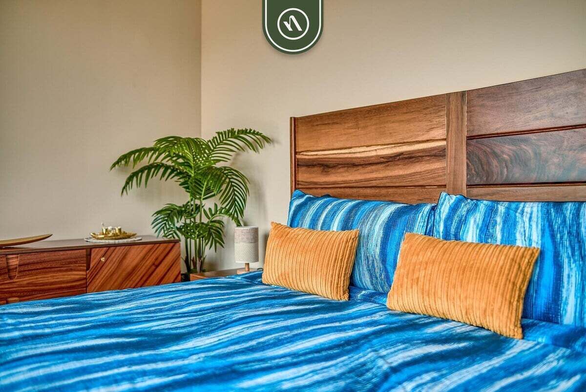 A bed with a blue striped bedspread and two orange pillows, a wooden headboard, a potted plant, and a wooden dresser.