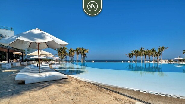 A poolside area with lounge chairs and umbrellas, palm trees, and a clear view of the ocean.