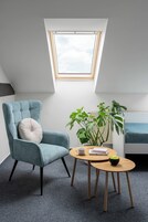 Una habitación con tragaluz, un sillón azul, una mesa redonda de madera, una planta en maceta y una cama con colcha azul.