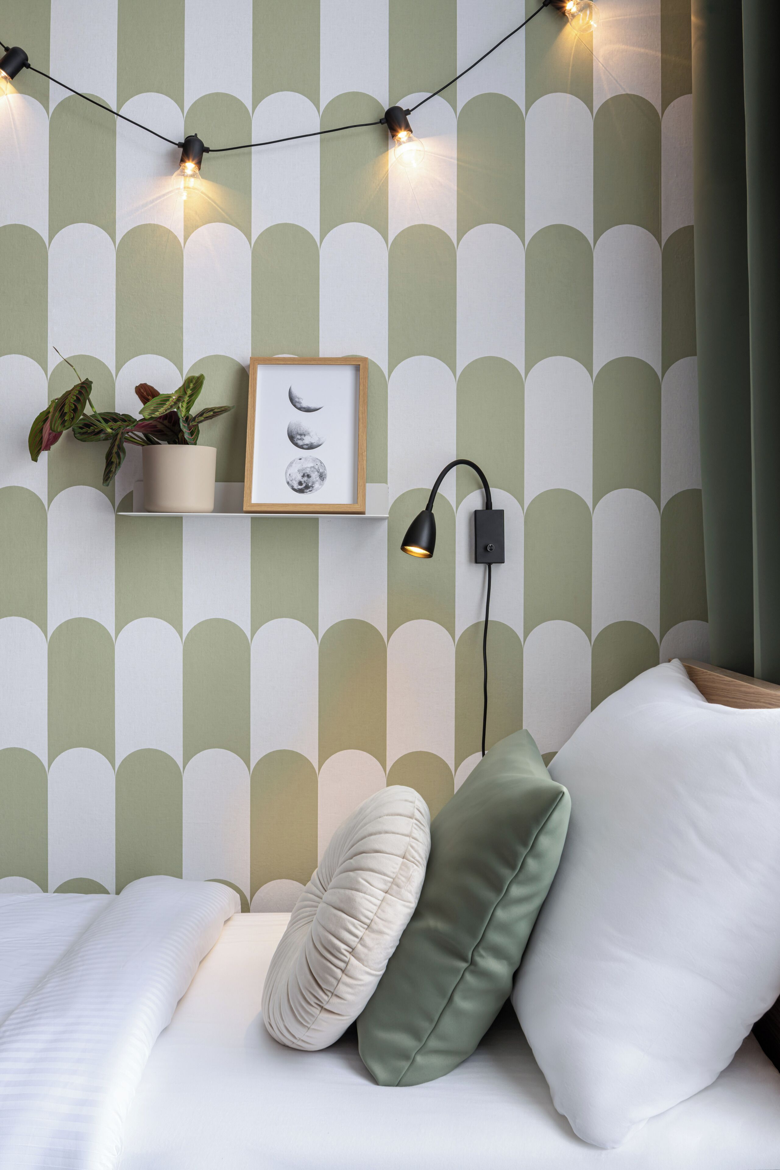 Un dormitorio con cama, almohadas, un estante en la pared con una planta y un cuadro enmarcado, y una lámpara de pared.