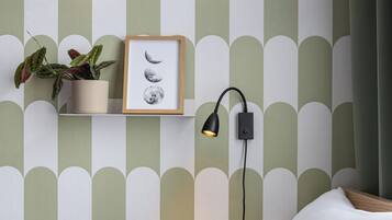Un dormitorio con cama, almohadas, un estante en la pared con una planta y un cuadro enmarcado, y una lámpara de pared.
