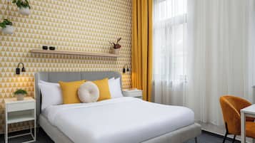 Ett modernt hotellrum med en vägg som har ett geometriskt mönster, en säng med vita sängkläder och gula detaljer, ett sängbord med en växt och en stol med ett mönstrat kuddöverdrag.