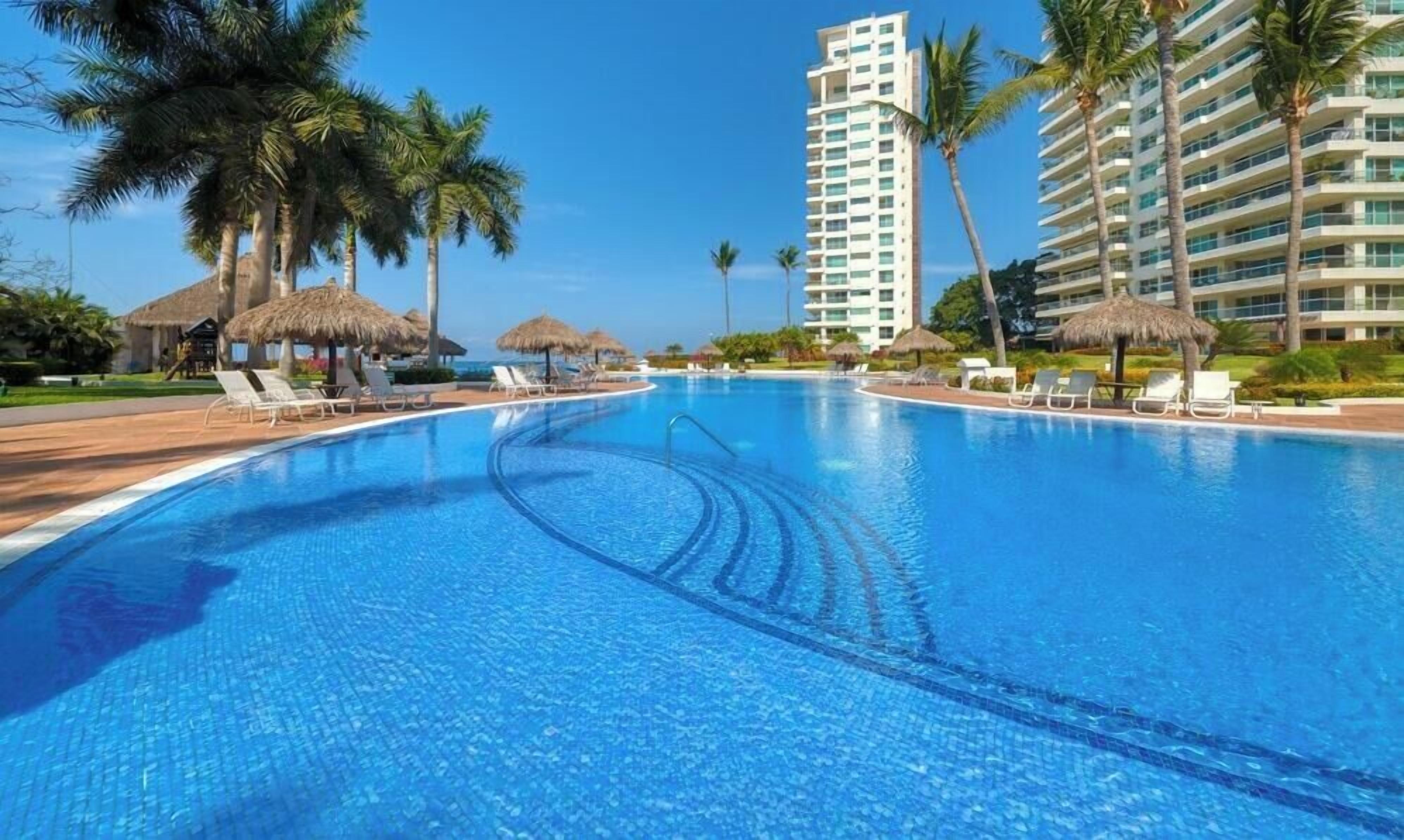 A swimming pool with lounge chairs, palm trees, and a tall hotel building.