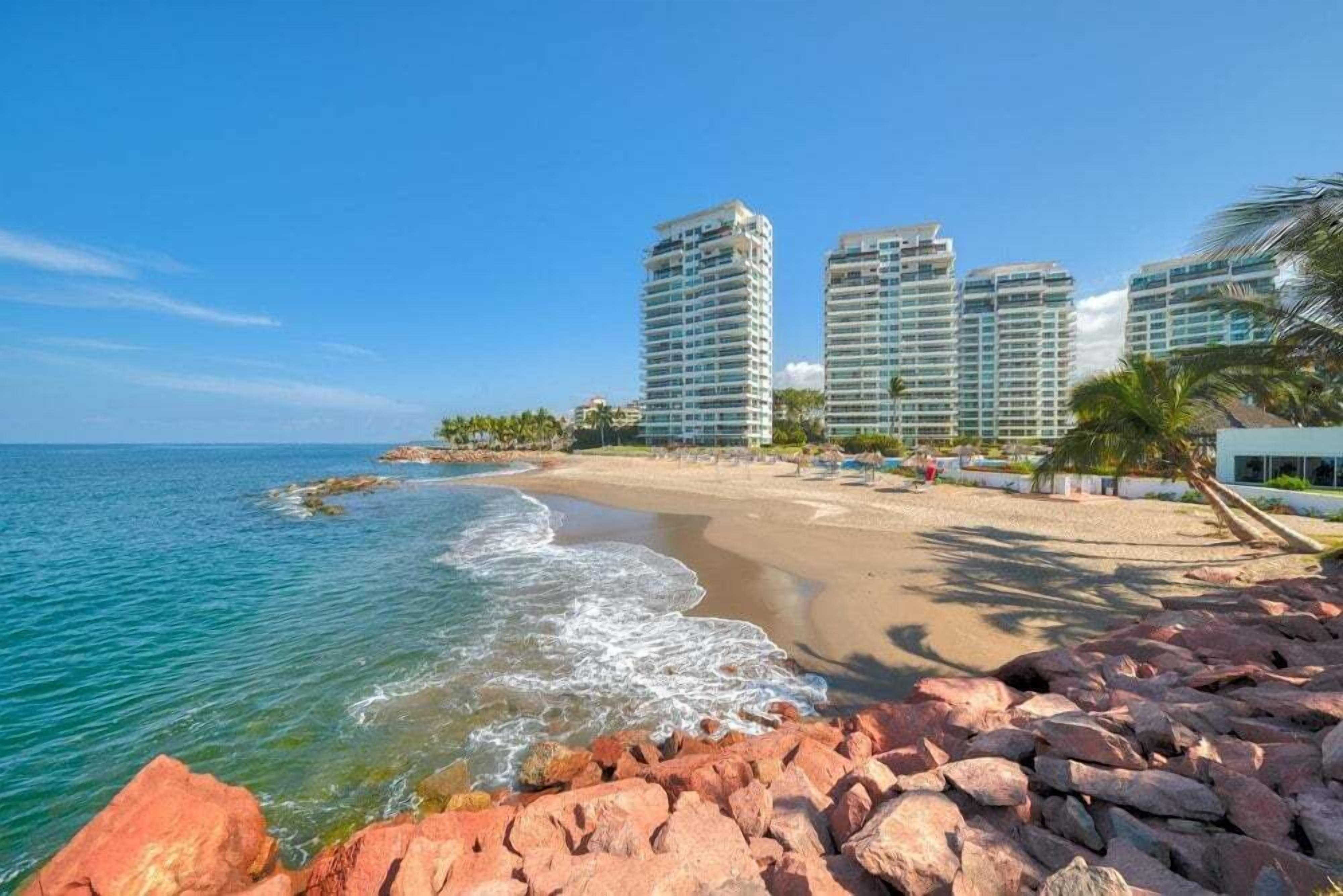 Una spiaggia con uno sfondo roccioso, mare cristallino e grattacieli sullo sfondo.