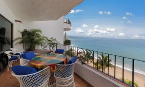 Ein Balkon mit Tisch und Stühlen, der Blick auf einen Strand mit dem Meer und einem klaren Himmel bietet.