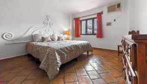 A bedroom with a patterned bedspread, a decorative headboard, a wooden dresser, and a window with red curtains.