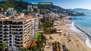 Un paysage urbain côtier avec une plage, un grand hôtel doté de nombreux balcons et une chaîne de montagnes en arrière-plan.