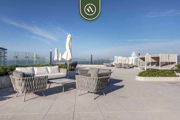 Una terraza en la azotea con mobiliario exterior, barandilla de vidrio y vistas al horizonte de la ciudad.
