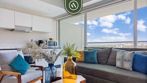 A modern living room with a sofa, a coffee table, and a chair. There is a view of the city through large windows. - New 2BR APT - Beach Access - Pool - GYM - Jacuzzi (Nuevo Vallarta)