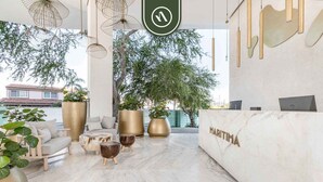 A reception area with a marble reception desk, seating area with sofas and chairs, large potted plants, and hanging pendant lights. - New 2BR APT - Beach Access - Pool - GYM - Jacuzzi (Nuevo Vallarta)