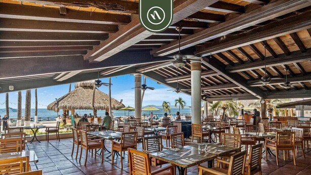 An outdoor dining area with wooden tables and chairs, a thatched roof, and a view of the beach and ocean. - Costa Sur Estudio 4 | Beachfront Studio w/ Pool & A/C (Puerto Vallarta)