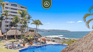 A resort pool area with a clear view of the ocean and mountains in the background, surrounded by palm trees and thatched-roof huts. - Costa Sur Estudio 4 | Beachfront Studio w/ Pool & A/C (Puerto Vallarta)