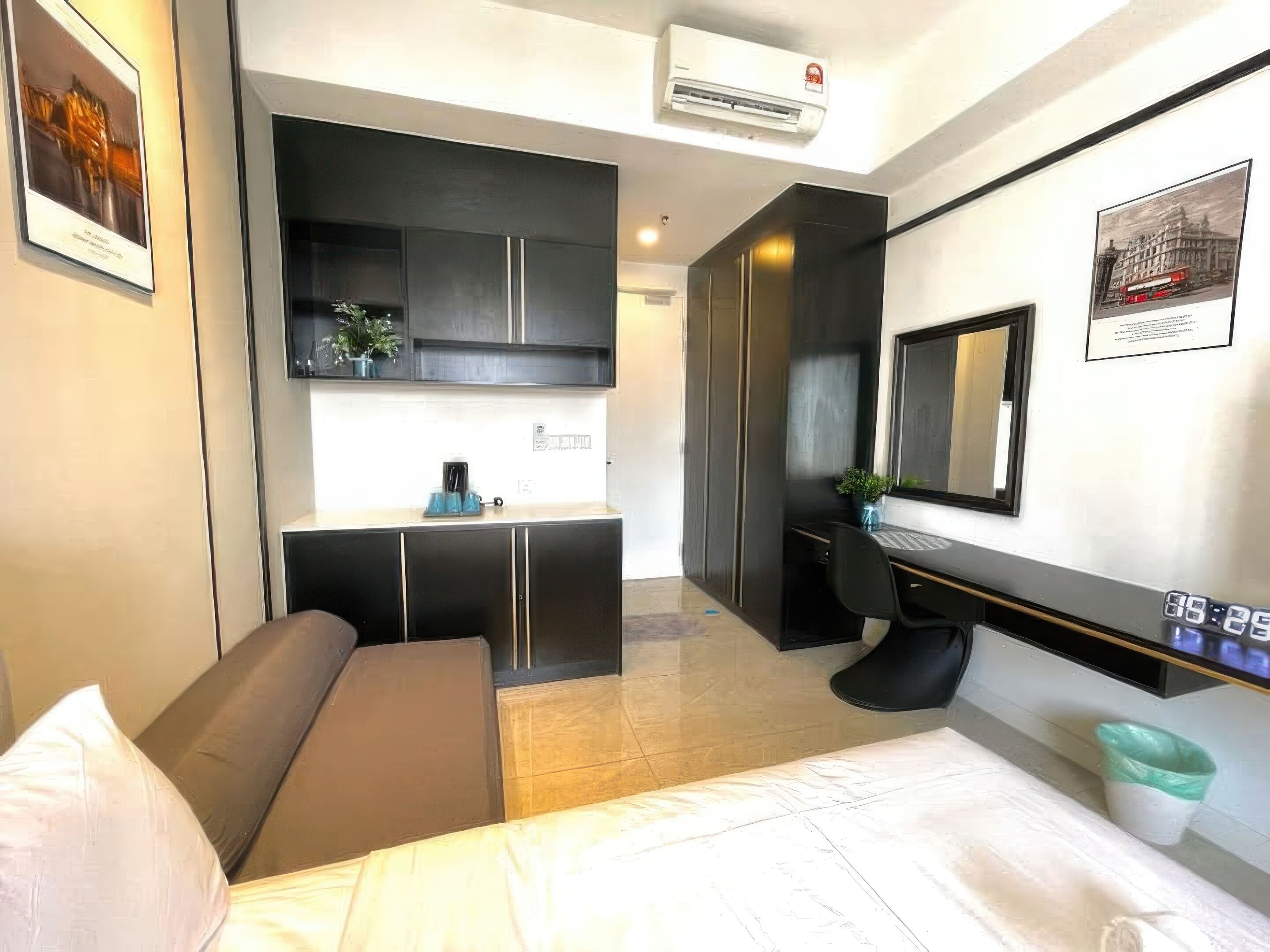 A compact hotel room with a bed, a desk, a chair, a kitchenette, and a large mirror.