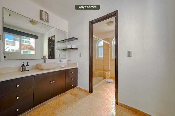 Une salle de bain dotée d’un grand miroir, d’un lavabo, de meubles sombres et d’un espace douche visible à travers une porte en verre.