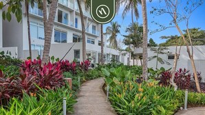 A pathway flanked by lush greenery and palm trees, leading to a modern building with balconies.