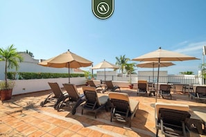 A rooftop terrace with lounge chairs and umbrellas, a clear sky, and a palm tree.