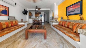 Ein Wohnzimmer mit Couchtisch, zwei Sofas mit gemusterten Kissen, Deckenventilator und einer Küche im Hintergrund.