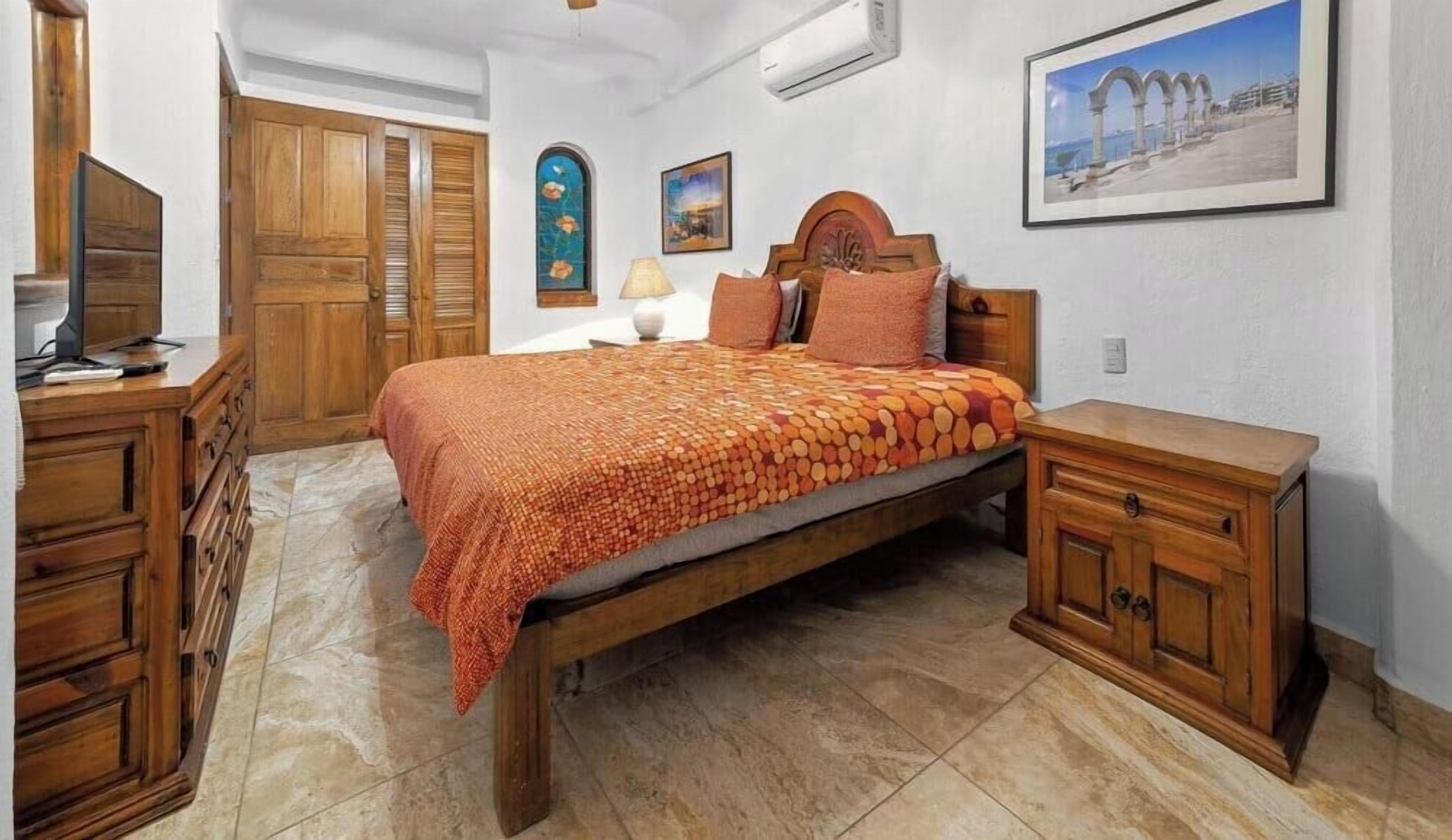 Een slaapkamer met een houten bed, een houten dressoir, een televisie, een nachtkastje en een ingelijst schilderij aan de muur.