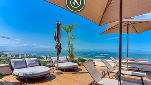 A deck with lounge chairs and a table, overlooking a scenic view of the sea and mountains. - New 1 BR Condo with Amazing Rooftop Pool - Jacuzzi (Bucerías)