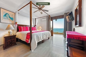 A four-poster bed with a ceiling fan, a bedside table with a lamp, a view of the ocean, and a balcony with a table and chairs.