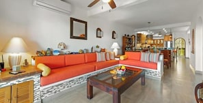 A spacious living room with a large orange sofa, a glass coffee table, and a kitchen in the background. - 603 Plaza Mar | 2BR/2BA Puerto Vallarta | Pool, Balcony (Puerto Vallarta)