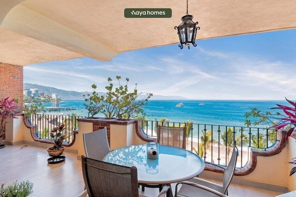 A balcony with a glass table and chairs, a hanging lantern, and a view of the ocean and distant cityscape.