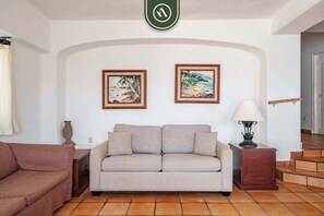 A living room with a beige sofa, two framed beach paintings, a lamp, and a staircase.