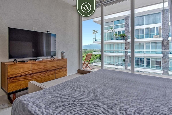 Une chambre d’hôtel moderne avec un grand lit, un téléviseur à écran plat posé sur une commode en bois, et un balcon offrant une vue sur un immeuble et des palmiers.