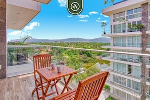 Un balcon avec une table et des chaises, donnant sur un jardin luxuriant et des montagnes lointaines.