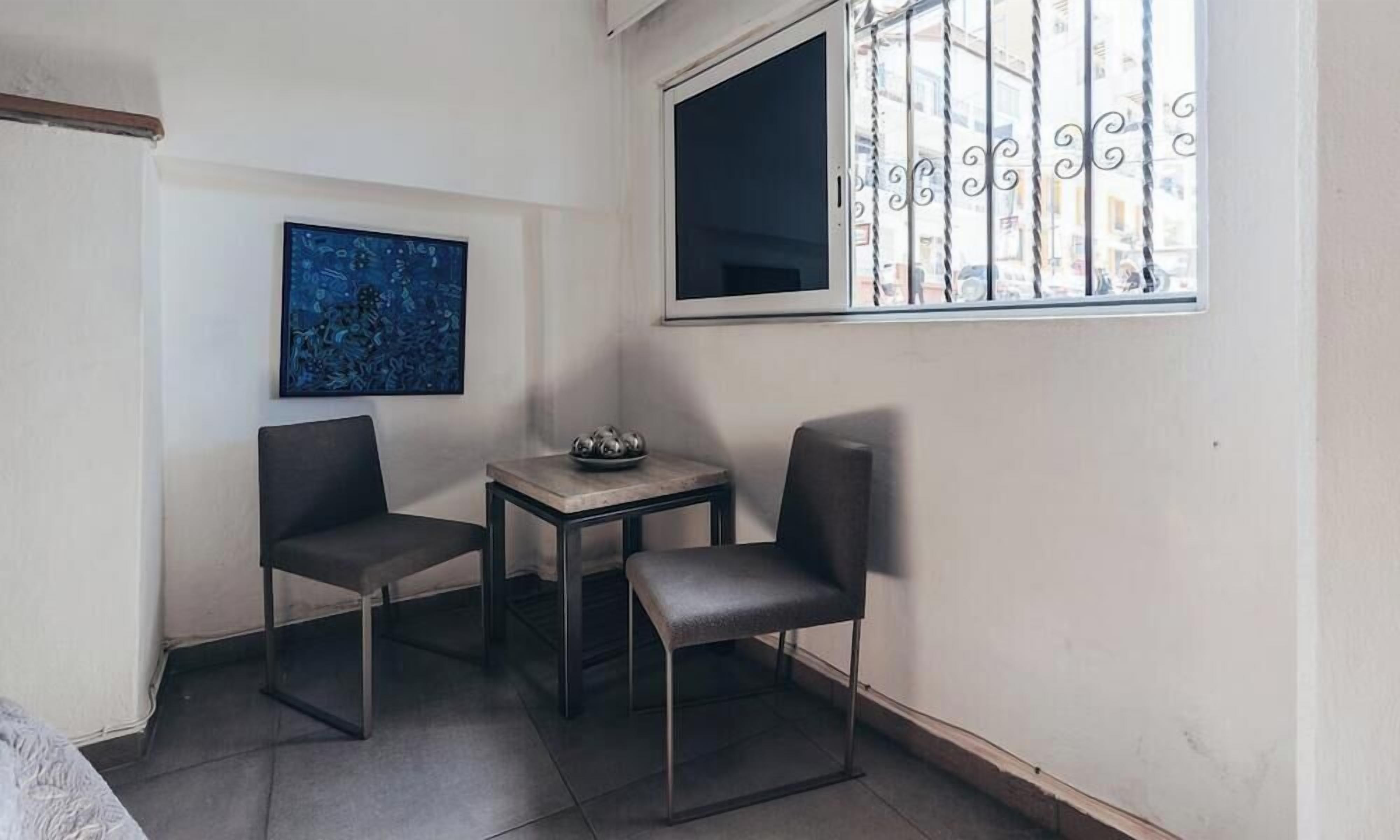 Une petite pièce, de taille modeste, comprenant deux chaises et une table, une fenêtre avec des barreaux en métal, et une œuvre d’art abstraite accrochée au mur.