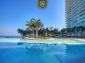 Ein großer, klarer Pool mit Palmen und Liegestühlen, umgeben von einem Hochhaus.