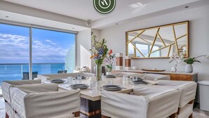 A modern dining area with a view of the ocean, featuring a large table set for six, a vase with flowers, and a decorative mirror on the wall.